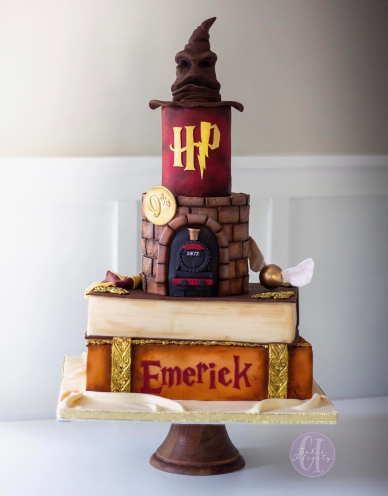 Harry Potter-themed tiered birthday cake for a 1st birthday celebration. The cake features stacked book layers, adorned with edible decorations resembling iconic Harry Potter elements, including a train, Golden Snitch, and Sorting Hat perched on top