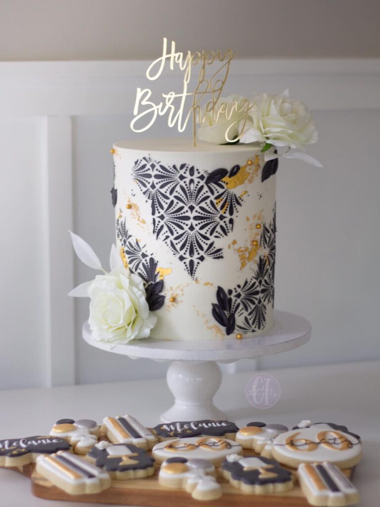 60th Birthday Celebration Cake: An elegant 8" double-barrel cake with white buttercream, adorned with sophisticated black stenciling. Custom gold acrylic decorations add a touch of personalized luxury to this tall and stylish birthday confection