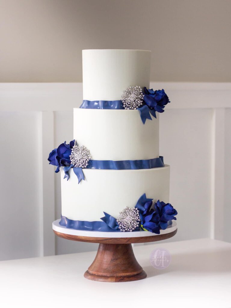 Traditional Navy & White Wedding Cake: A classic 3-tier confection in sizes 10"-8"-6", adorned with buttercream and elegant fondant ribbon. The cake features refined brooch details, adding a touch of sophistication to this timeless navy and white wedding centerpiece