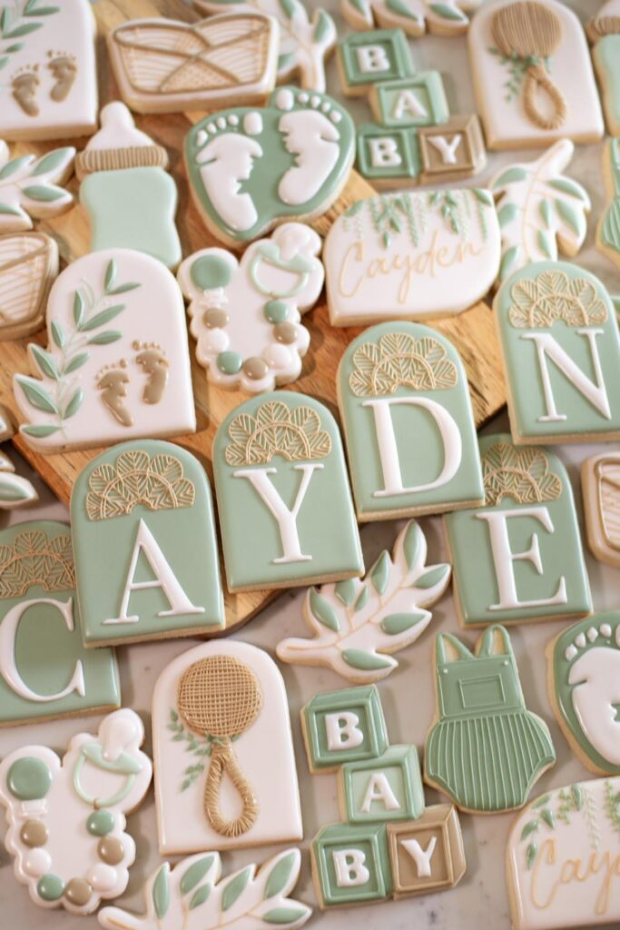 Cayden's Baby Shower Cookies: A charming assortment with a boho feel, featuring wicker detailing and a soothing palette of sage green and beige. The cookies include sweet details like baby feet, a bassinet, and personalized treats, creating a delightful and personalized touch for Cayden's baby shower celebration