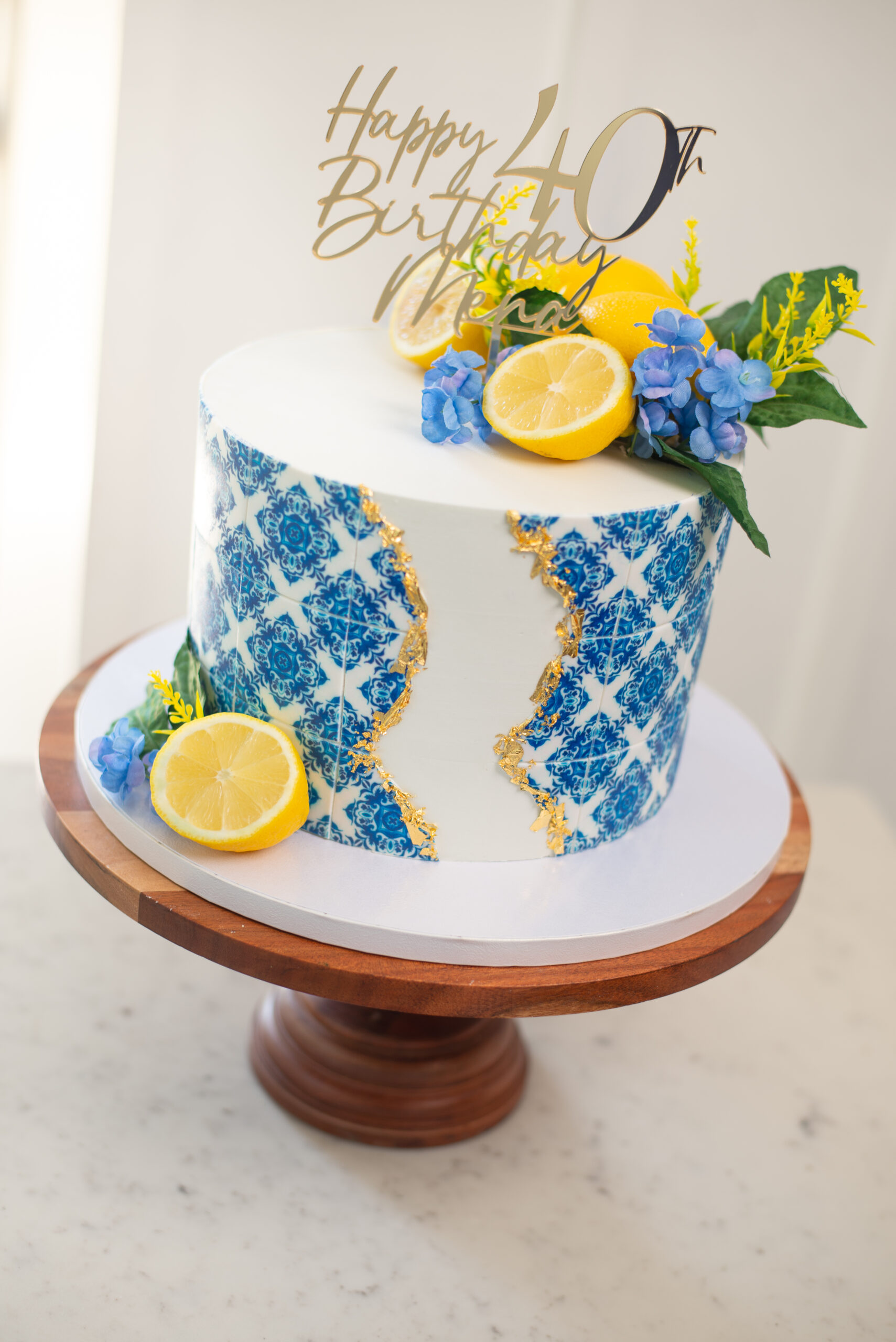 40th Birthday Cake: An 8" Italian-inspired masterpiece adorned with a blue and white tiled detail around the side. The cake is elegantly decorated with lemons and greenery, featuring a custom gold acrylic topper. The fresh and clean design follows a Navy, yellow, and white color scheme, creating a stunning centerpiece for the 40th birthday celebration