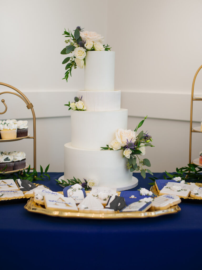 4-Tier Modern Wedding Cake: An elegant masterpiece featuring a clean, all-white design. One tier showcases fluted fondant detailing, adding a touch of sophistication. Adorned with stunning florals provided by the florist, this cake creates a beautiful and modern centerpiece for a wedding celebration